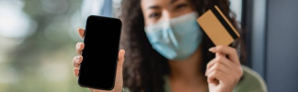 Beyaz ekranlı akıllı telefon ve bulanık arka planda tıbbi maskeli Afrikalı Amerikalı bir kadının elinde kredi kartı. - Fotoğraf, Görsel