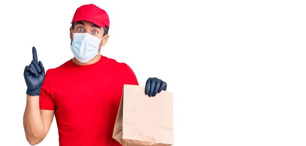 Fiatal spanyol férfi ételt szállít, kovid-19 biztonsági maszkot visel, papírzacskót tart, meglepve egy ötlettel vagy kérdéssel, ami ujjal mutogat boldog arccal.  - Fotó, kép