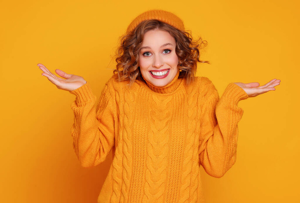 Gelukkig jong vrouwtje in trendy trui en hoed schouders ophalen en glimlachen voor de camera tegen gele backgroun - Foto, afbeelding