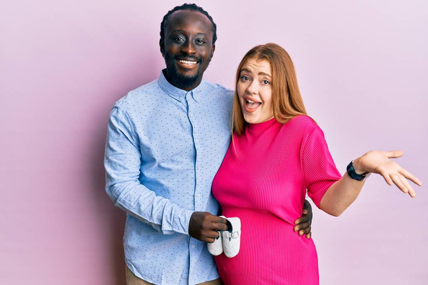 Joven pareja interracial esperando un bebé sosteniendo zapatos celebrando el logro con sonrisa feliz y expresión ganadora con la mano levantada  - Foto, Imagen