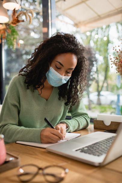 african american woman in medical mask writing in notebook near laptop in cafe - Photo, Image