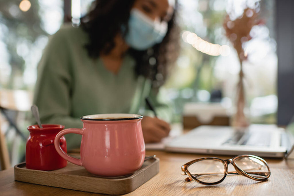 чашка кофе возле сахарной чашки и африканская американка в медицинской маске на размытом фоне  - Фото, изображение