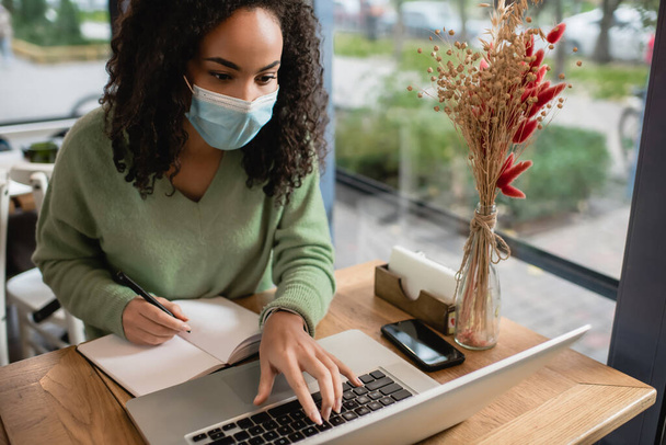 mujer afroamericana en máscara médica utilizando el ordenador portátil cerca de teléfono inteligente y portátil en la mesa  - Foto, imagen