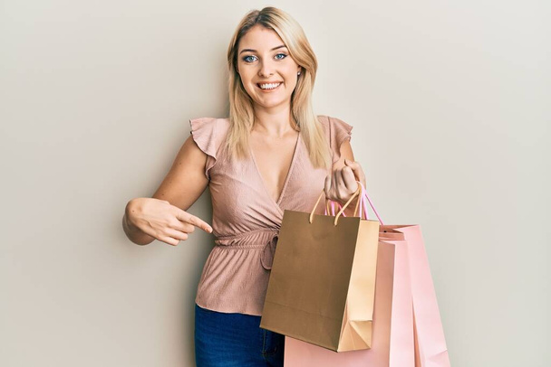 Joven mujer caucásica sosteniendo bolsas de compras sonriendo feliz señalando con la mano y el dedo  - Foto, Imagen