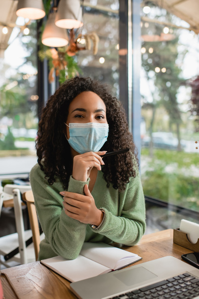 Afrikanerin mit medizinischer Maske blickt in die Kamera in der Nähe von Gadgets und Notizbuch auf dem Tisch im Café - Foto, Bild