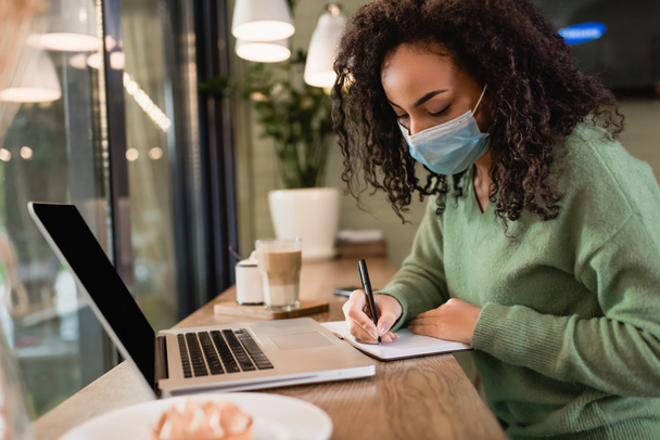 african american woman in medical mask writing in notebook near laptop with blank screen on table  - Foto, afbeelding