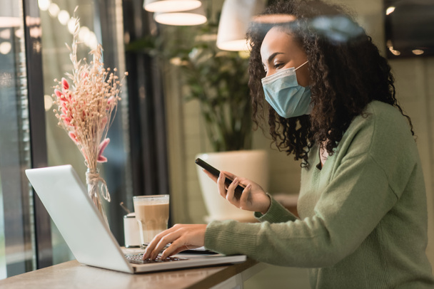 curly african american freelancer in medical mask holding smartphone near laptop and glass of latte in cafe  - Fotografie, Obrázek