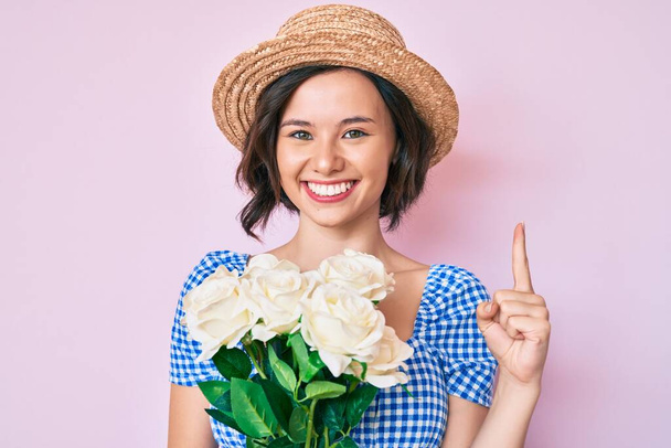 Menina bonita nova usando chapéu de verão segurando flores sorrindo com uma ideia ou pergunta apontando o dedo com o rosto feliz, número um  - Foto, Imagem
