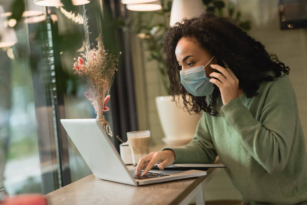 donna afro-americana in maschera medica parlare su smartphone mentre si utilizza laptop vicino notebook sul tavolo - Foto, immagini