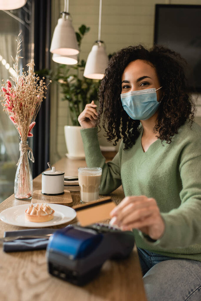 mujer afroamericana en máscara médica pagando por tarta y café en cafetería - Foto, imagen