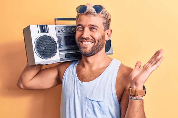 Joven hombre rubio guapo con barba usando gafas de sol escuchando música usando boombox celebrando el logro con sonrisa feliz y expresión ganadora con la mano levantada - Foto, Imagen
