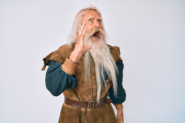 Vieil homme âgé aux cheveux gris et à la barbe longue portant un costume traditionnel viking criant et criant haut et fort, la main sur la bouche. concept de communication.  - Photo, image