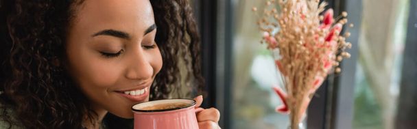 mujer afroamericana complacida sosteniendo taza rosa con café en la cafetería, pancarta - Foto, imagen