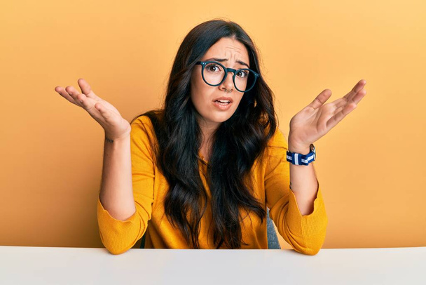 Beautiful brunette young woman wearing glasses and casual clothes sitting on the table clueless and confused expression with arms and hands raised. doubt concept.  - Photo, Image