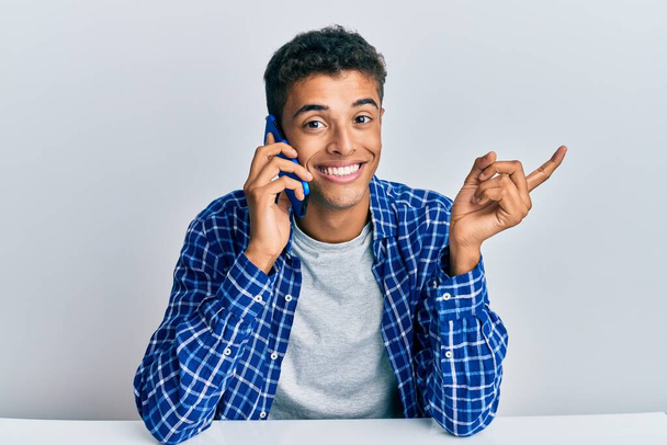 Young handsome african american man having conversation talking on the smartphone smiling happy pointing with hand and finger to the side  - Photo, Image