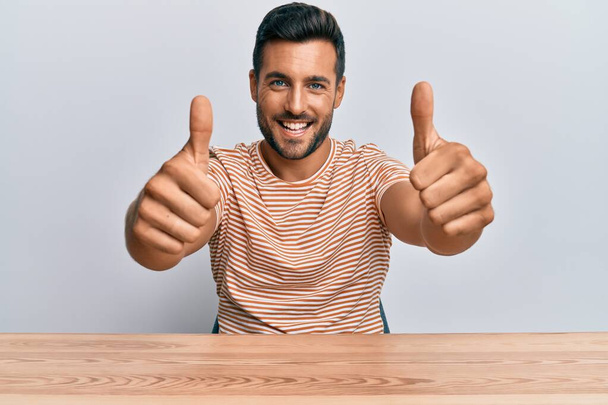 Hombre hispano guapo usando ropa casual sentado en la mesa aprobando hacer gesto positivo con la mano, pulgares arriba sonriendo y feliz por el éxito. gesto ganador.  - Foto, imagen