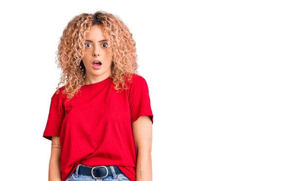 Young blonde woman with curly hair wearing casual red tshirt in shock face, looking skeptical and sarcastic, surprised with open mouth  - Photo, Image