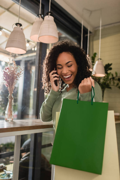 alegre afroamericana mujer hablando en smartphone y sosteniendo bolsas de compras en la cafetería - Foto, Imagen