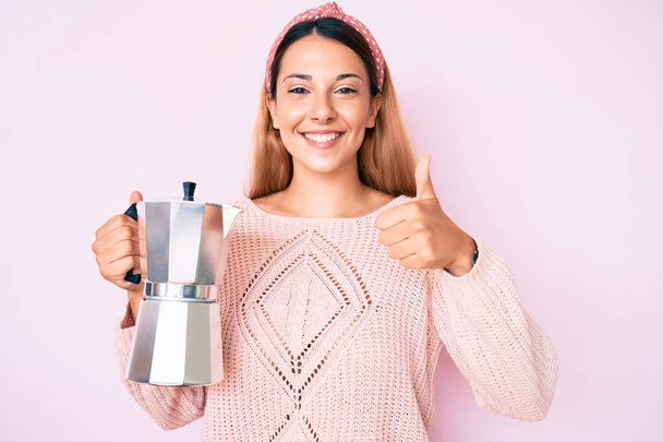 Jonge brunette vrouw met Italiaanse koffiezetapparaat glimlachen gelukkig en positief, duim omhoog doen uitstekend en goedkeuring teken  - Foto, afbeelding