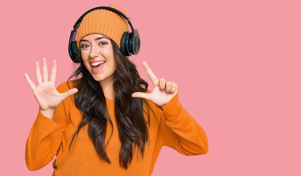 Όμορφη μελαχρινή νεαρή γυναίκα ακούγοντας μουσική χρησιμοποιώντας ακουστικά που δείχνουν και δείχνουν προς τα πάνω με τα δάχτυλα νούμερο επτά, ενώ χαμογελά αυτοπεποίθηση και χαρούμενος.  - Φωτογραφία, εικόνα