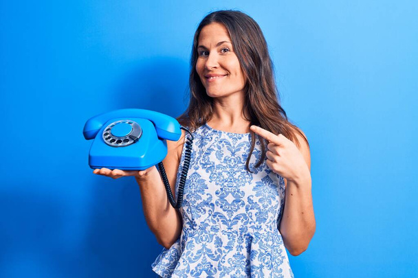 Молодая красивая брюнетка с винтажным телефоном на изолированном синем фоне улыбается счастливо указывая рукой и пальцем - Фото, изображение