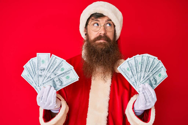 Beau jeune homme roux à longue barbe portant santa claus costume tenant tas de dollars souriant regardant vers le côté et regardant loin en pensant.  - Photo, image