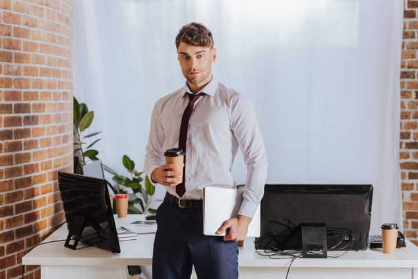 Joven hombre de negocios sosteniendo café para llevar y portátil cerca de computadoras en la oficina  - Foto, imagen