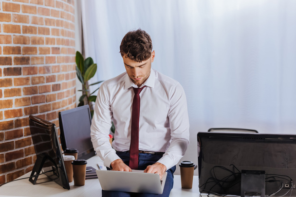 Businessman in formal wear using laptop near coffee to go and computers in office  - Foto, Imagem