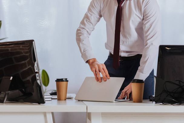 Обрізаний вид бізнесмена, що тримає ноутбук біля витягування кави та комп'ютерів в офісі
  - Фото, зображення