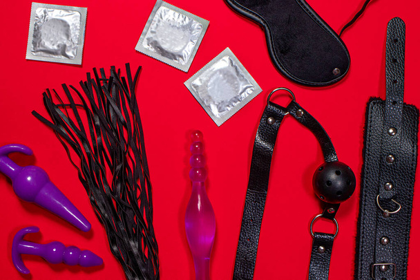 Yetişkinler için seks oyuncakları ve kırmızı arka planda prezervatifler. Mantar, kırbaç, öğürme. Korunan seks, doğum kontrolü ve BDSM. - Fotoğraf, Görsel