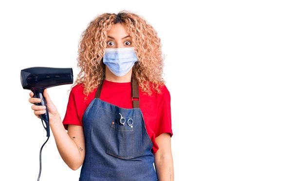 Young blonde woman with curly hair holding dryer blow wearing safety mask for coranvirus scared and amazed with open mouth for surprise, disbelief face  - Foto, imagen