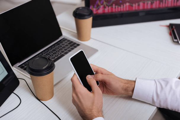 Vista recortada del hombre de negocios usando un teléfono inteligente con pantalla en blanco cerca de café para llevar y computadoras en un fondo borroso  - Foto, Imagen