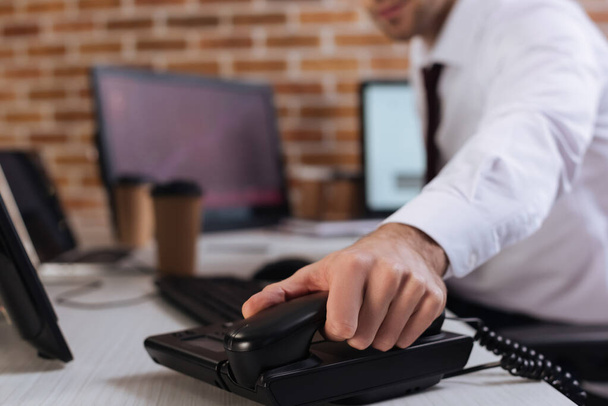 Vista cortada do empresário segurando telefone auscultador perto de computadores em fundo embaçado  - Foto, Imagem