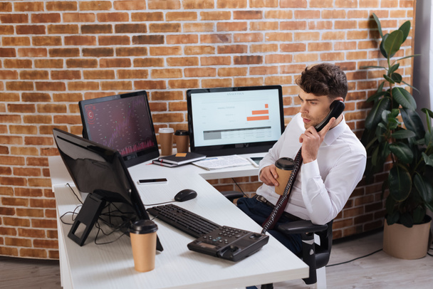 Бізнесмен тримає винос кави і дивиться на комп'ютери під час розмови по телефону
  - Фото, зображення