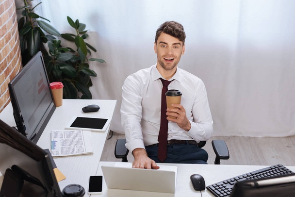 Uomo d'affari sorridente che tiene il caffè per avvicinarsi ai dispositivi e al giornale in ufficio  - Foto, immagini