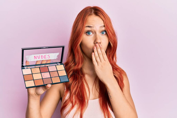 Young redhead woman holding makeup nudes covering mouth with hand, shocked and afraid for mistake. surprised expression  - Photo, Image