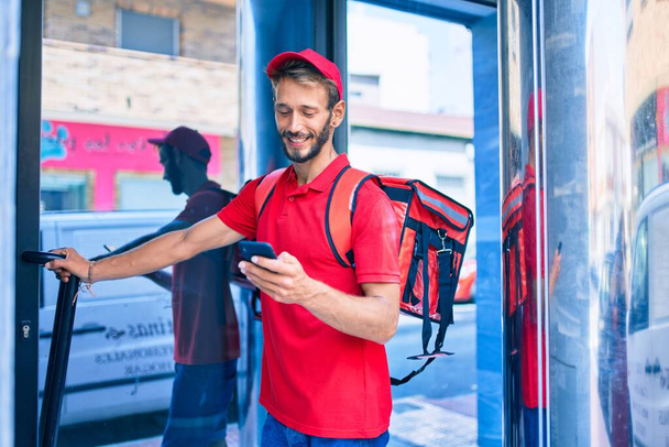 Кавказький доставник у червоній формі і рюкзак доставки посміхається на відкритому повітрі за допомогою смартфона - Фото, зображення