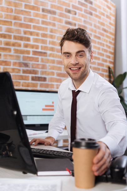 Smiling businessman taking coffee to go near computer on blurred foreground  - Photo, Image