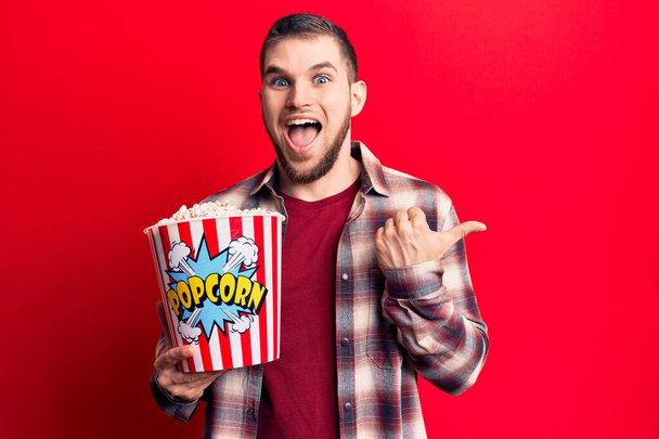 Young handsome man eating popcorn pointing thumb up to the side smiling happy with open mouth  - Photo, Image