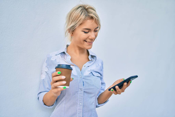 Młoda blondynka uśmiecha się szczęśliwy za pomocą smartfona i picia zabrać kawę w mieście. - Zdjęcie, obraz
