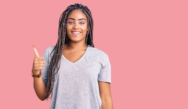 Νεαρή Αφροαμερικανή γυναίκα με κοτσίδες που φοράει καθημερινά ρούχα χαμογελώντας χαρούμενη και θετική, με τον αντίχειρα να κάνει εξαιρετική δουλειά και το σήμα έγκρισης  - Φωτογραφία, εικόνα