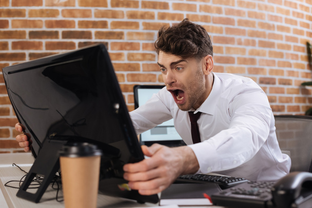 Empresario loco sosteniendo monitor de computadora en primer plano borroso mientras revisa el mercado financiero en la oficina  - Foto, imagen