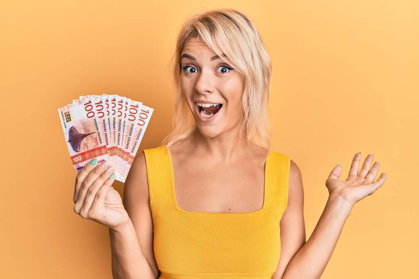 Mladá blondýnka drží norské koruny bankovky slaví úspěch se šťastným úsměvem a vítězným výrazem se zvednutou rukou  - Fotografie, Obrázek