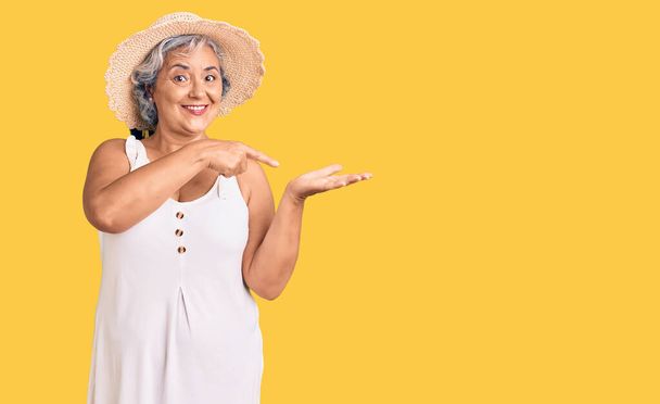 夏の帽子と夏の服を身に着けている灰色の髪のシニア女性は、手で提示し、指で指している間、カメラに驚いて笑顔.  - 写真・画像