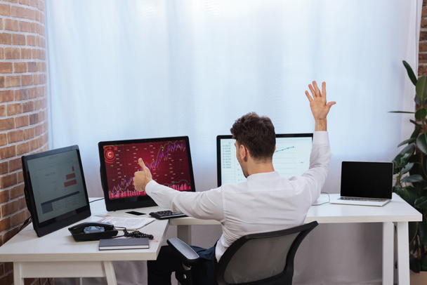 Обратный вид бизнесмена, показывающего большой палец вверх возле компьютеров с графиками финансового рынка на мониторах  - Фото, изображение