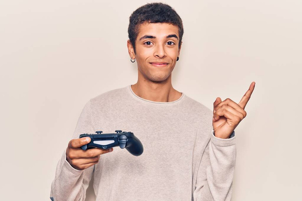 Молодий афроамериканський чоловік грає в відеогру, тримаючи контролер усміхненим щасливим вказуючи рукою і пальцем в бік
  - Фото, зображення