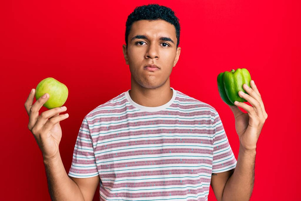 Mladý arabský muž držící zelené jablko a pepř skeptický a nervózní, zamračený kvůli problému. negativní osoba.  - Fotografie, Obrázek