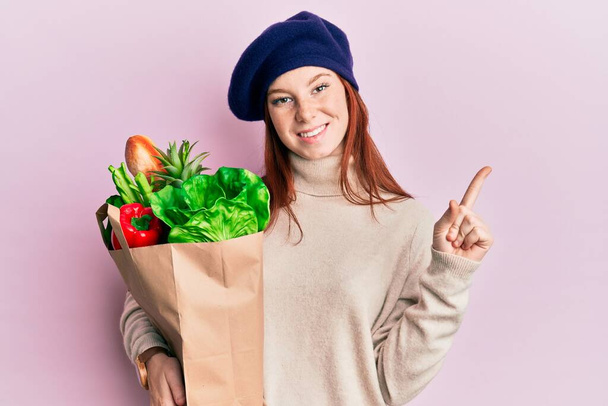 Young red head girl holding paper bag with bread and groceries smiling happy pointing with hand and finger to the side  - Foto, Imagen
