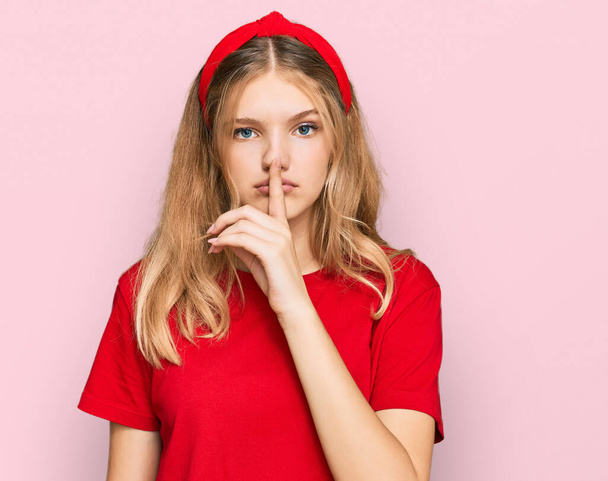 Schöne junge Kaukasierin in lässigem rotem T-Shirt, die mit dem Finger auf den Lippen um Ruhe bittet. Schweigen und geheimes Konzept.  - Foto, Bild