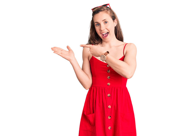 Νεαρή όμορφη ξανθιά γυναίκα φορώντας casual φόρεμα κατάπληκτη και χαμογελώντας στην κάμερα, ενώ παρουσιάζει με το χέρι και δείχνοντας με το δάχτυλο.  - Φωτογραφία, εικόνα